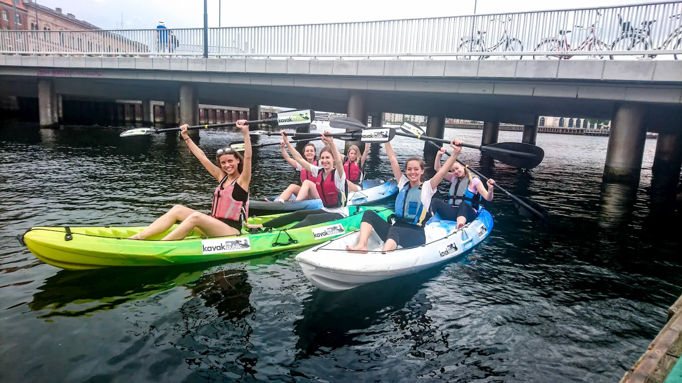 Group of people in double-kayaks in front of bridge